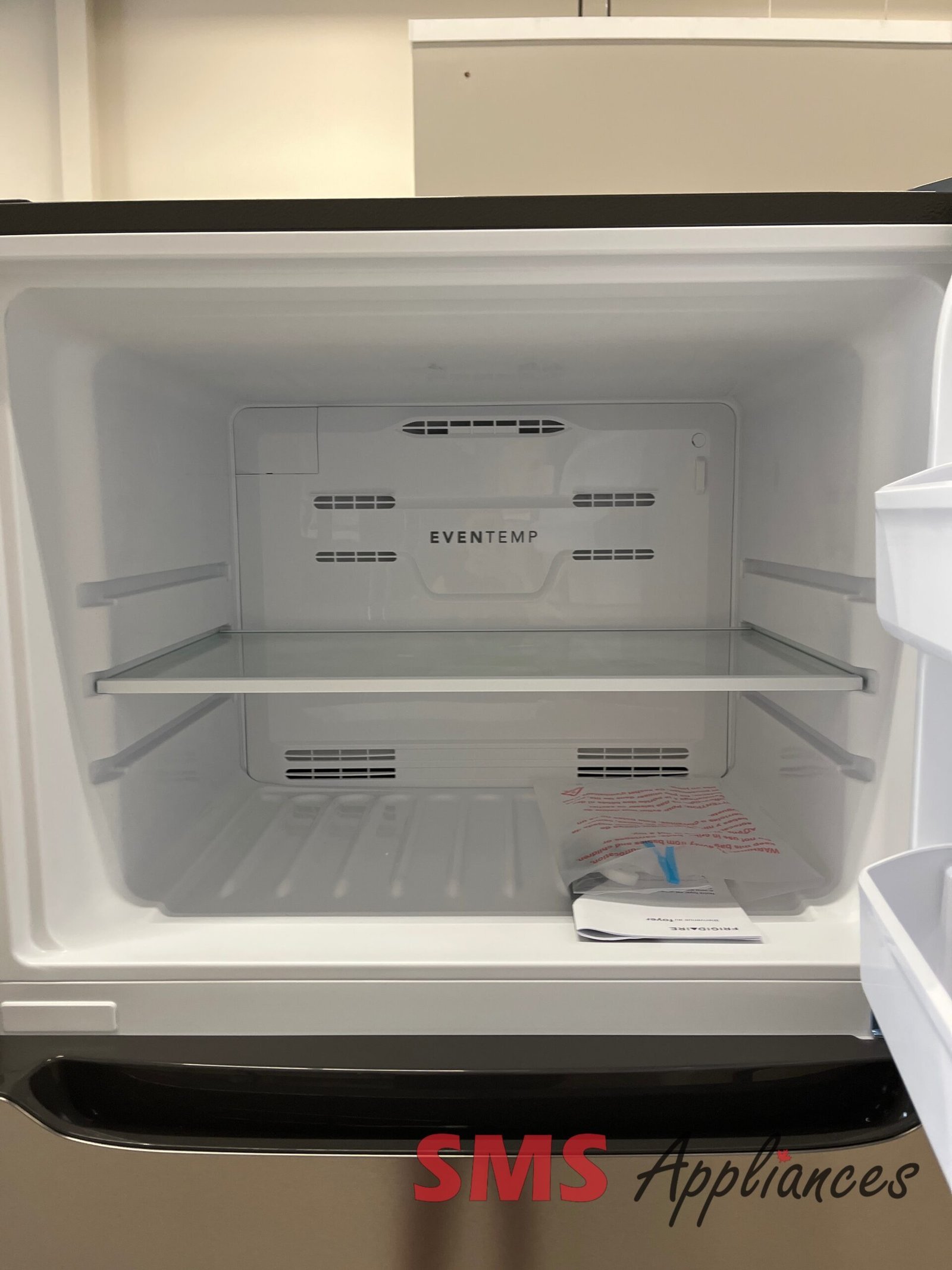 Open-Box-Fridges-FRTE1835AV-3-3-scaled-1.jpg Open refrigerator with two metal shelves and an empty interior; a few papers lie on the bottom shelf, and the SMS Appliances logo is visible on the exterior front.