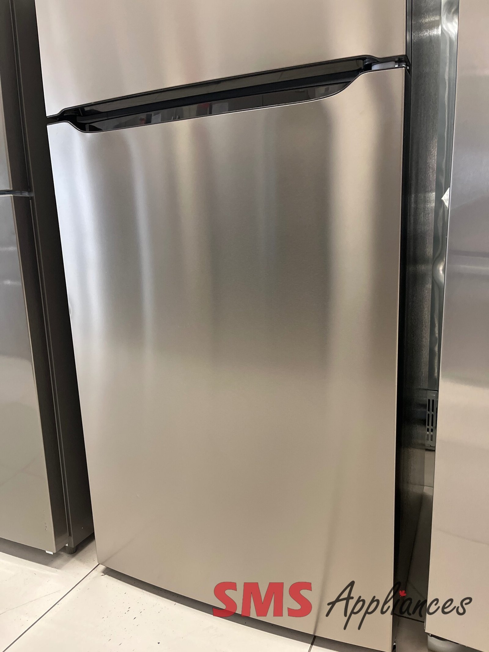 Open-Box-Fridges-FRTE1835AV-7-2-scaled-1.jpg Stainless steel refrigerator with a bottom freezer drawer and integrated curved handle in a showroom setting, brand signage is visible at the base.