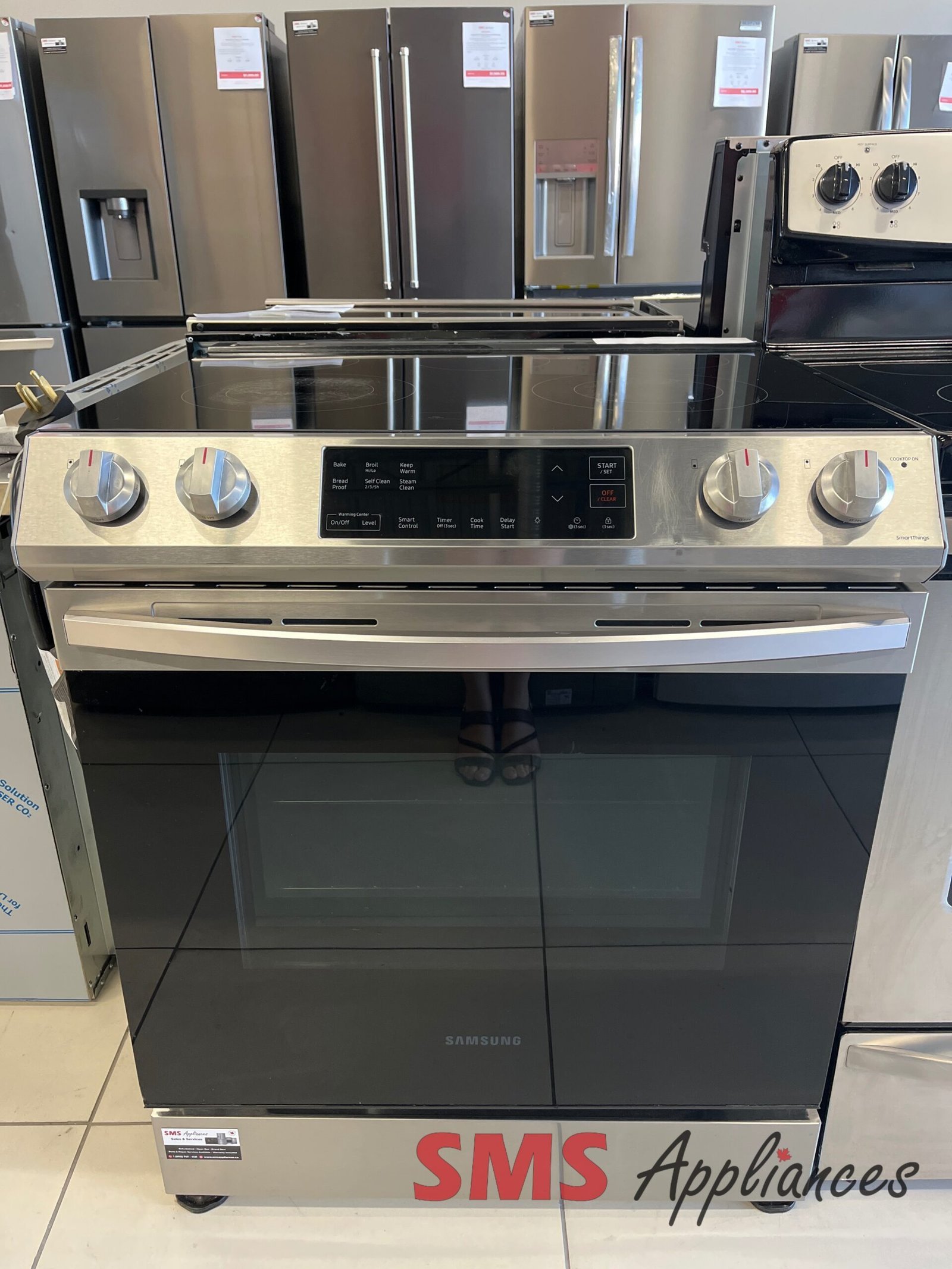 Refurbished-Stoves-Ranges-NE63T8111SS-1-scaled-1.jpg Samsung stainless steel range with glass cooktop on display in an appliance store, surrounded by multiple refrigerators in the background.