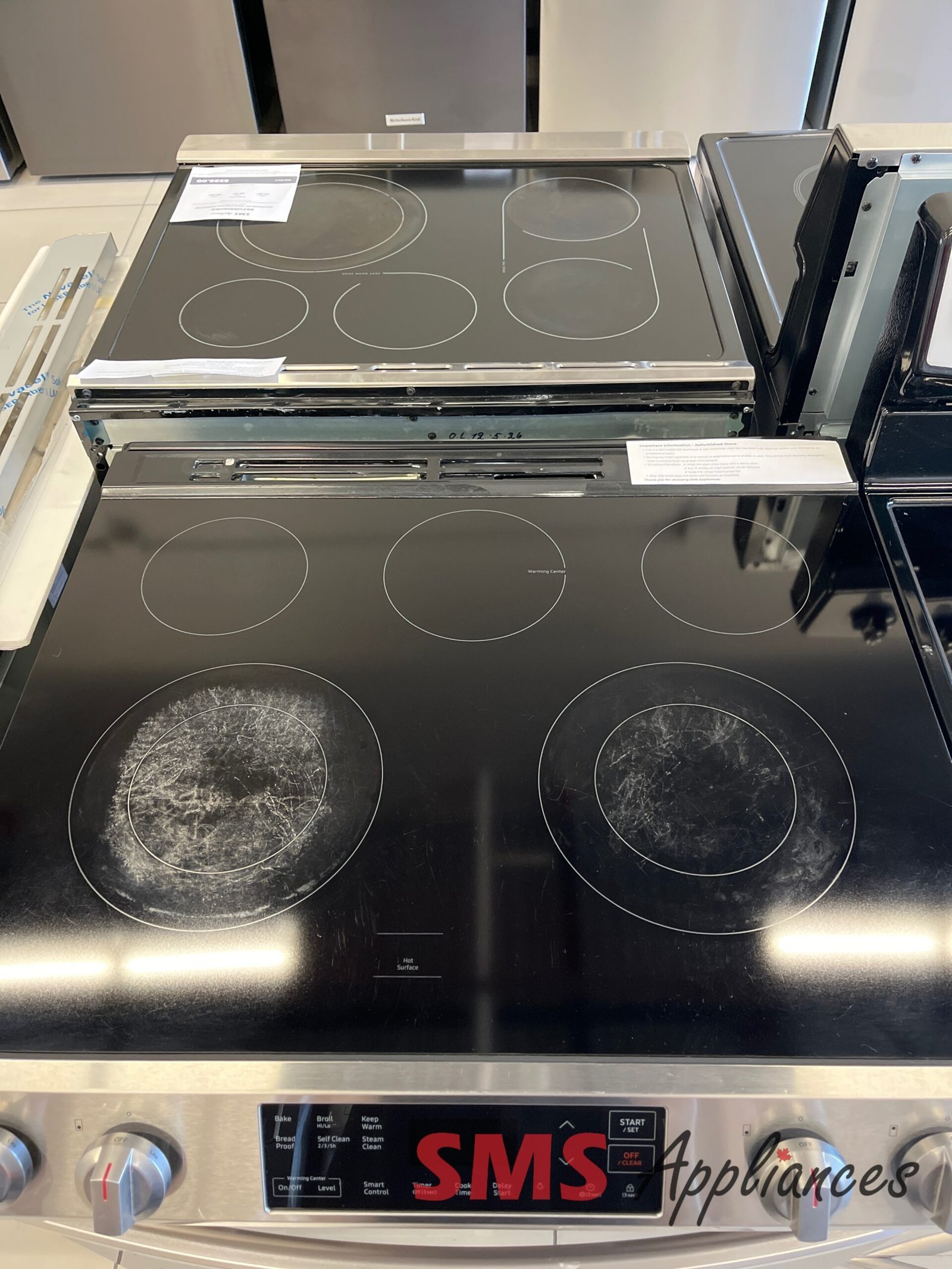 Refurbished-Stoves-Ranges-NE63T8111SS-3-scaled-1.jpg Black glass-ceramic induction cooktop with multiple circular burners and stainless trim, viewed in a showroom setting.
